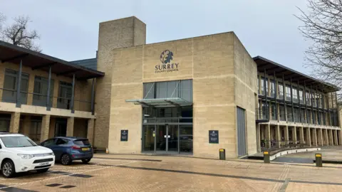 The entrance to the Surrey County Council offices at Woodhatch. The sign is above the door.

Two cars are also parked outside on the pavement