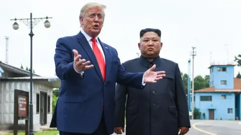 Getty Images Standing on a road in the Joint Security Area, Trump gestures with his hands as he speaks, wearing a navy suit and red tie. Beside him stands Kim Jong Un, wearing a black button-up suit, looking solemn with his hands by his side.