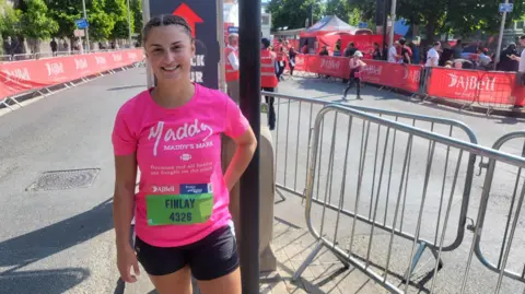 Finlay O'Ragen is wearing a pink shirt that says Maddy's Mark on it. She is standing on the road that is cordoned off for the runners. She is smiling.