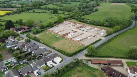 John Fairhall/BBC A drone image of the developed site, this time taken from further back and from a different angle but showing the same scene. It is a field that has been largely concreted over and has several caravans, both static and mobile, on it. Fencing is being installed around the perimeter of the site, which is flanked by residential housing and other fields.