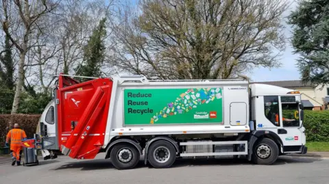 A Biffa recycling lorry is parked on a residential street beside wheelie bins. The vehicle displays a large "Reduce Reuse Recycle" sign and branding from Tandridge District Council and Biffa, with collection staff visible nearby.