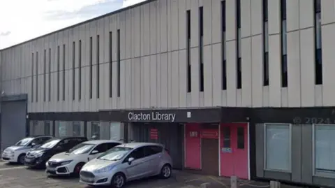 Clacton Library. The building has a red door and several cars parked outside it. The building grey. It has a sign that says Clacton Library at the entrance.