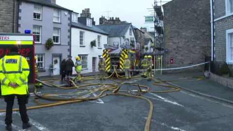 BBC View down a street in which stand firefighters and two fire engines, with a tangle of fire hoses on the ground. A police officer stands on the left of the scene and a firefighter is talking to a man and a woman dressed in dark clothing.