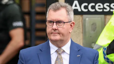 Jeffrey Donaldson. He has short grey hair, wearing glasses, a blue blazer, white shirt, blue and yellow tie, with a fish pin on his lapel.