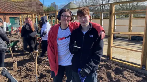 BBC Morgan has a spade in their hand while Roo has her arm around their shoulder. There is a building between them. There is mud on the ground. 