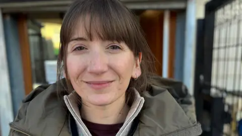 BBC Lucy Cox stands outside a building. She is wearing a jumper and winter jacket and has her brown hair tied back.