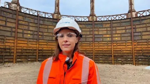 Andrew Turner/BBC Nadia McInnes is wearing a white hard hat and orange high visibility jacket. She has brown hair, which is at shoulder length, and she is wearing safety glasses. She is standing within the tank are of the Great Yarmouth gasometer, which has had the lid removed, and exposed the inside structure of the rusting cast iron tank which shows ribs and vertical rails.