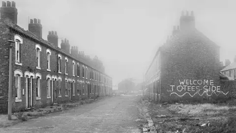Peter Benson A black and white photograph of an empty derelict terraced street with weeds growing on the pavements. On one side is ironic graffiti painted on the gable end of a house which reads Welcome to Teesside.