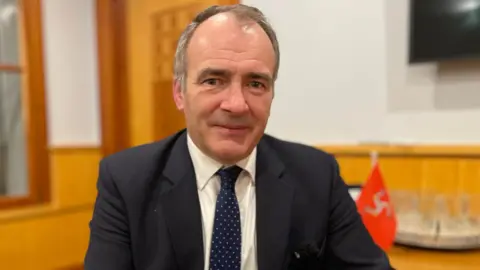 Alfred Cannan is wearing a black suit with a white shirt and dark spotty tie. He his dark greying hair and is clean shaven. He is sitting in his office panelled with. pine wood; a small red Manx flag can be seen on a desk behind him.