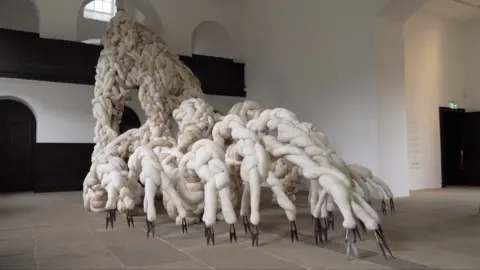 A sculpture with tendrils, made out of wool and horse hair in an empty room with white walls