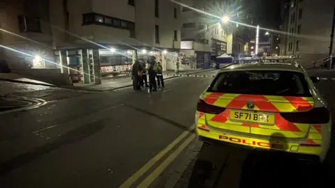 A police car parked on the pavement in Cowgate in Edinburgh. There is a double yellow line on the road. In front of the fence there is a police officer talking to several people dressed in dark clothes.