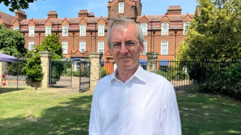 Tom Williams/BBC Sir Laurie Bristow smiles at the camera while standing in the grounds of the college, wearing a white open necked shirt and glasses. He is squinting slightly in the sun.