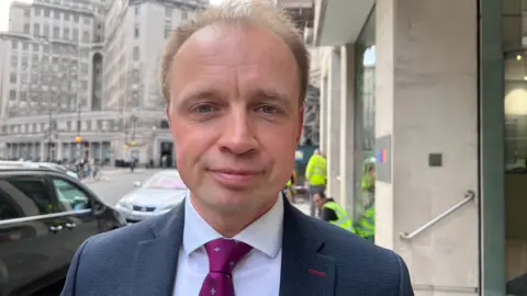 Southampton Itchen MP Darren Paffey stands outside an office building. He has short fair hair and is wearing a dark blue suit and a purple tie.