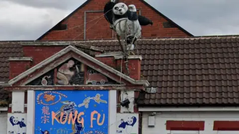 An ornamental figure of a black and white panda kicking one leg in the air. The figure is on the roof of a building which has a blue sign on the wall which says 'Kung Fu'.