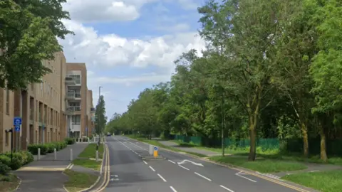 A Google StreetView image showing part of Grahame Park Way in Edgware, with a modern block of flats on the left and a thick row of trees on the right, with a pedestrian crossing in the middle of the road