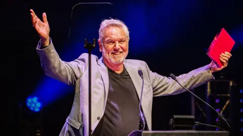 Andrew Billington Brian Conley stands behind a lectern with two small microphones holding both hands in the air and smiling at the audience. He wears a silver coloured suit jacket over a grey V-necked top and has short hair with smart stubble.