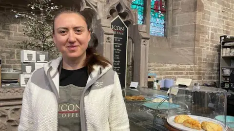 A woman with brown hair and wearing a grey and white jacket stands in a church. A stone wall and trays of pastries can be seen behind her, as well as signs for the coffee shop.