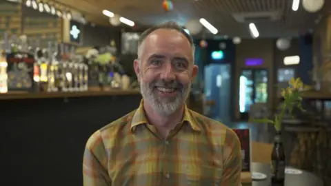 Man in the foreground wearing a yellow and green checked shirt, seated indoors and facing the camera. The setting is a bar or pub, with a row of beer taps and bottles visible behind the person on the left. Tables and chairs are positioned in the background, and a small vase containing yellow flowers sits on a table to the right. Overhead ceiling lights and hanging decorations are visible throughout the venue. No bags, bins or markers are visible.