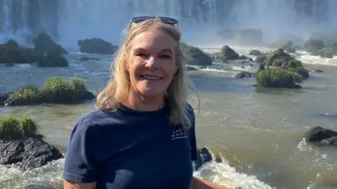 Jackie Stacey A woman with blonde hair and a blue top stands in front of a waterfall