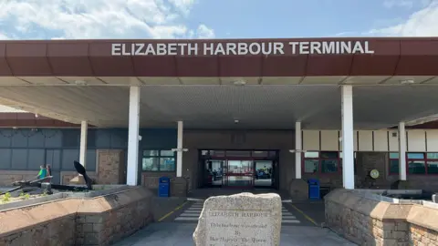 A picture of the front of Elizabeth harbour terminal. It is a large building with a glass entrance and a stone in front of it.