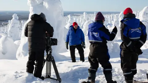 David standing in an Arctic landscape wearing a red hat, thermals and gloves. There are three people closest to the camera who are part of a filming crew.