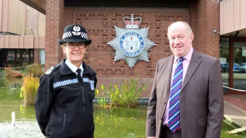Suffolk Constabulary Rachel Kearton and Tim Passmore standing outside Suffolk Constabulary's HQ. Rachel is on the left and wearing a navy police hat with blue and white trim, a navy police zip-up jacked over a white shift and glasses. Tim is wearing a light pinky brown jacket over a pink shirt and tie. Behind them is a pond and a brick wall with the Suffolk Police log on it.