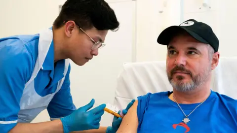 PA Media A nurse is on the left of the picture with a needle, inserting it into Steve's arm. Steve is sat on a chair and is wearing a blue top and a black baseball hat. He is wearing a metal chain round his neck with a small jigsaw piece pendant. 