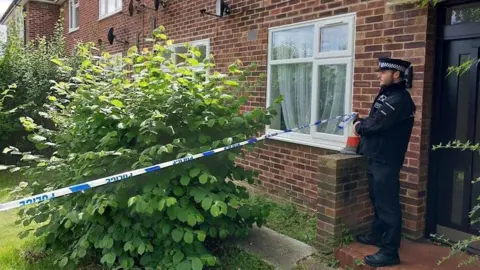 Essex Police A police officer stood outside the front door of a terraced red-brick house. He is next to a police cordon that has been placed around the front garden, which contains a large green bush.