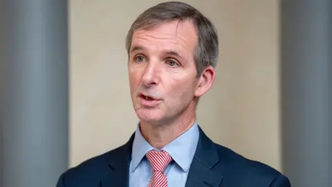 PA Media Liam McArthur, who has short grey hair, speaks while turned slightly to his right. He is wearing a dark blue suit, light blue shirt and red and white stripy tie. 
