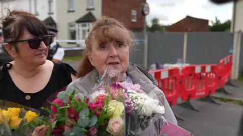 Elsie Dot Stancombe's aunt Jean Stevenson clutches a bouquet of flowers as she speaks to reporters close to the police cordon in Southport. She is comforted by a woman in dark glasses. 