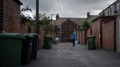 Back lane in Horden in County Durham. The houses are red brick, with back yards and a scattering of greenery. There's a wooden telegraph pole and green wheeled bins lined up against the back walls. A teenaged boy, wearing a royal blue t-shirt and dark trousers, is walking away.