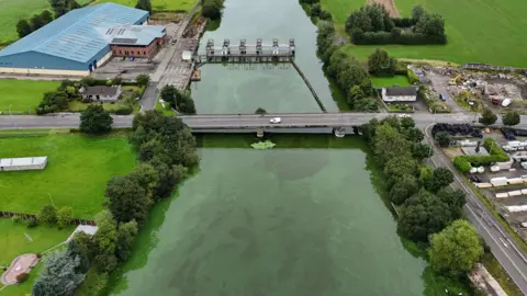 PA Media A drone shot of a river covered in green sludge. In the centre of a photo is a bridge crossing the river. On each side of the river are green fields and deciduous trees. A road runs along the right-hand bank.