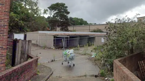 A set of derelict garages which are falling apart. A road leads up to the garages, and an abandoned shopping trolley sits in the middle of the road.