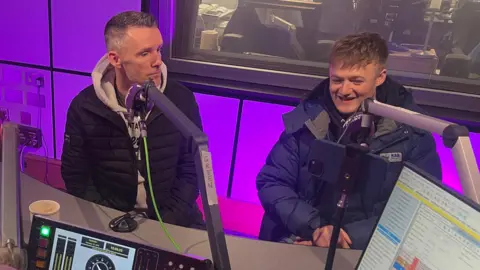 Two men sit in a radio studio in front of microphones. The man on the left has short dark hair, a grey hoodie and black puffer jacket. The man on the right has short fair hair and is wearing a thick navy coat. A computer screen, digital clock and sound equipment sit on the desk in front of them. The studio is lit with purple lighting and has a large glass panel leading to a production area with office chairs and speakers.