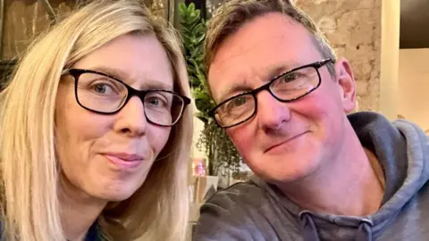 Family photo A woman with long blond hair and black framed glasses and a man with short light brown hair and black framed jacket in a grey top in a room with brick walls and a plant behind them