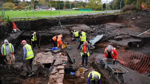 Stuart Walker Photography About a dozen people are wearing high-vis jackets and working on the dig site, either digging with shovels or moving wheelbarrows. Beyond is a grassy field and a red-bricked building.