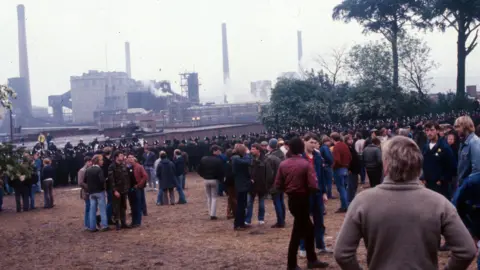 A still from BBC video footage taken on the day, which shows hundreds of men in everyday 80s clothing spread out across a grass lawn, with the wall of police officers stood in front of the industrial plant, its chimneys rising into the air.
