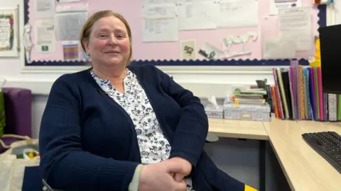 Maria smiles at the camera as she sits in a chair by her desk. She has a blue cardigan with a white shirt with floral patterns on.  She has light brown hair tied into a pony tail.