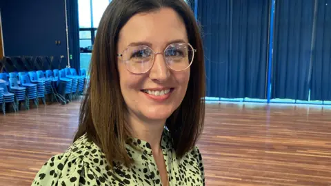 Zoe is standing in a rehearsal space. She is wearing a black and green top and glasses. There are blue curtains drawn in the background and a stack of plastic chairs in the corner.