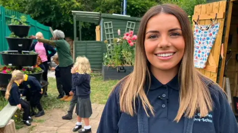 A young woman with long blonde hair smiles in a garden. She is wearing a navy blue polo shirt and fleece. To her left four small children can be seen standing and sitting on a bench. Behind are green painting sheds and a fence. Two adult volunteers stand chatting next to planters. Green railings line the left hand side of the site.