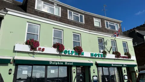 Exterior of the Dolphin Hotel in Jersey which has green walls, several windows with planters containing pink flowers in them and a sign saying Dolphin Hotel bar and restaurant in green and red letter.