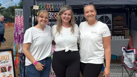 The image shows three women wearing white t-shirts standing outside a stall at the West Show.