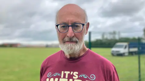 Tony Forster looking at the camera with a glum expression. He is wearing glasses and a dark red T-shirt. Behind him is the partially built green metal fence and a van on a field. 