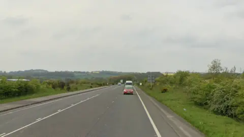 A red car travelling along Sprockhoevel Way an A road with green bushy verges.