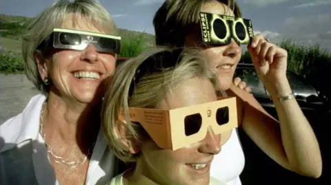 PA/Barry Batchelor A woman on the left wearing a white blouse, smiles while wearing her eclipse glasses. Her two daughters are both wearing the glasses which look to have a cardboard outer and black lenses. Behind them is a grassy verge. 