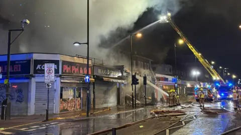 Two firefighters on a yellow turntable ladder spray water onto the fire from above. A row of shops, with homes set back from and above the shops, can be seen below along with a large plume of smoke