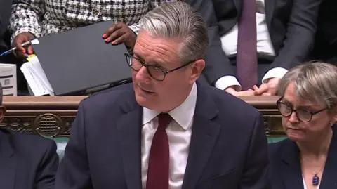 Parliament TV Sir Keir Starmer standing up in parliament. He is at the despatch box and has quiffed grey hair slicked to one side, glasses and a dark blazer and red tie over a white shirt.
