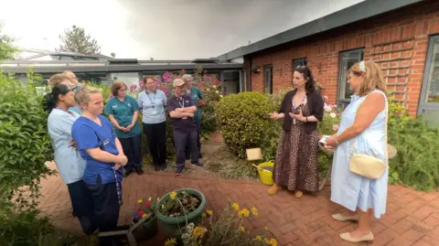 Sobell House Hospice staff gathered around in their inpatient unit garden as part of the time capsule ceremony. There are many flowers around. It is an overcast day.