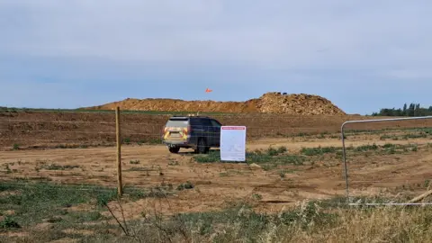 LDRS A large pile of dirt in the middle of a field. There is a black car parked near it and gates surrounding the site. 