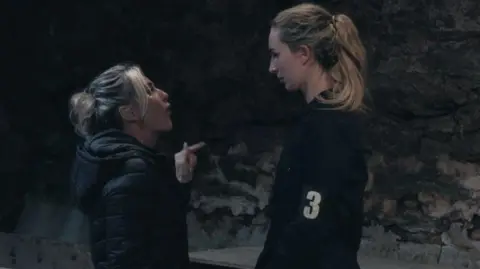 Channel 4 Michaella McCollum being interrogated as part of the final episode of Celebrity SAS. A small, blonde instructor wearing a black puffed coat points her finger at McCollum in a dark, stone wall room. McCollum stands on the right looking down. She is wearing a dark jacket with 3 on an armband. She has her long blonde hair tied back.